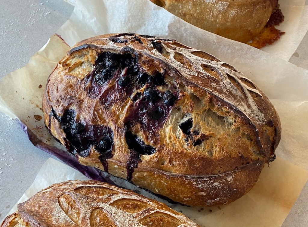 Artisan sourdough loaf loaded with whole wild blueberries and lemon zest, showing caramelized berry bursts and blistered crust